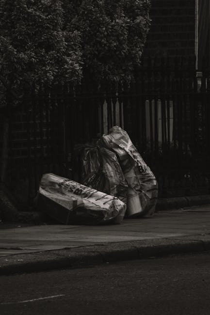 A group of three large, black plastic garbage bags filled with waste items are placed on the pavement beside a wooden fence, with a dense leafy bush partly visible behind it. The bags appear crumpled and tightly filled, with some partially torn or baggy material revealing creases in the plastic. They are positioned in a line, leaning slightly against each other, on a street surface that looks dark and smooth. The scene is illuminated by natural light, casting soft shadows on the sidewalk and highlighting the texture of the bags and nearby fencing. The wooden fence behind the waste is weathered and dark, with vertical slats, and extends across the background, suggesting a residential or private property. The surrounding environment indicates a typical outdoor urban setting where private waste disposal or rubbish collection might be arranged separate from council services, which relates to integrated rubbish removal and on-site clearance services that Waste Clearance Dulwich offers.