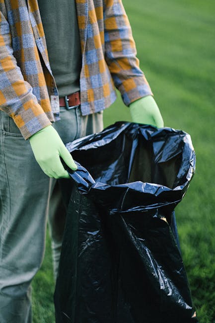 A person standing outdoors on a grassy area, wearing a multicoloured plaid shirt over a green t-shirt, with grey trousers and a brown belt. The individual is holding open a large black rubbish bag that is made from a durable plastic material with a slightly glossy finish. The person's hands are protected with light green gloves, and they are in the process of preparing the bag for waste collection, which could relate to private rubbish removal or on-site clearance services provided by companies such as Waste Clearance Dulwich. The background features a well-maintained lawn with no distinct structures or objects visible, emphasizing the outdoor environment suitable for waste collection activities. The lighting is natural, suggesting a clear day, and the scene reflects a typical waste disposal action associated with independent or non-local-authority rubbish handling practices, aligned with professional waste management services.