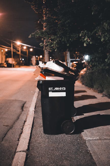 A black wheeled rubbish bin with the label 'ST. JOHN'S' is positioned on the pavement next to a curbstone, filled with various discarded waste items, including cardboard boxes, plastic containers, and paper. The bin is situated in an outdoor urban setting during nighttime, illuminated by streetlights which cast soft, warm glows and create shadows on the ground. In the background, there are trees, power lines, and a row of streetlights extending into the distance, suggesting a residential or mixed-use area. The surrounding environment appears calm, with no pedestrians or vehicle activity visible nearby. The scene emphasizes the importance of proper waste management through independent collection services that may be used as an alternative to public waste disposal, fitting within the context of rubbish removal options available in urban environments, often provided by companies like Waste Clearance Dulwich.