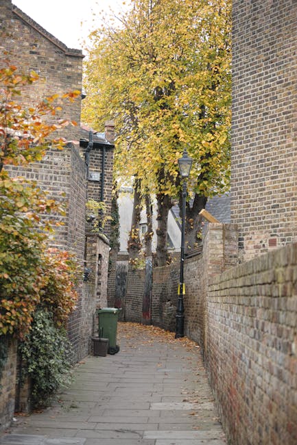 A narrow outdoor alleyway paved with concrete slabs, flanked by historic brick walls on both sides, with some bricks showing signs of weathering and age. In the foreground on the left, a green wheelie bin is positioned against the wall, while a small black trash container is nearby on the pavement. A tall, slender street lamp with a black finish and traditional design extends above the alley, casting subtle natural light onto the scene. Further into the background, a large tree with vibrant yellow and orange autumn leaves dominates the scene, its branches spreading over the alley and partially obscuring the buildings behind it. The alley appears to be a quiet, residential or historic area typical of urban environments, where private waste removal or on-site clearance may be conducted away from public-facing streets. The scene is well-lit with natural daylight, highlighting the textures of the brickwork, foliage, and paving stones, consistent with a setting where independent waste collection services like Waste Clearance Dulwich might operate to handle local rubbish removal in such community environments.