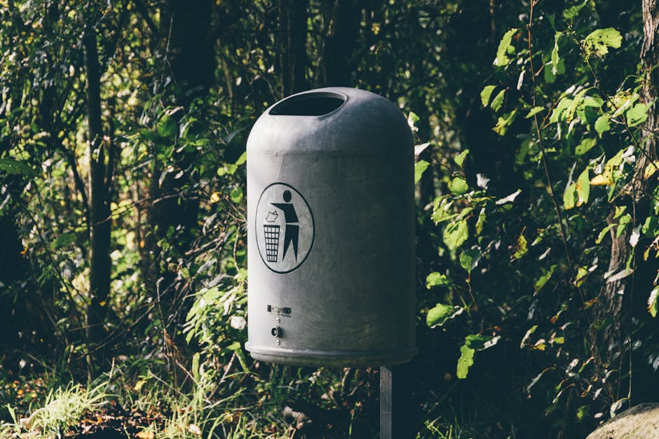 A standalone cylindrical metal refuse bin with a rounded top and an open slot on the upper side for trash disposal, positioned outdoors amidst dense green foliage and bushes. The bin is silver-grey, with a smooth, metallic finish, and a black symbol on the front depicting a person disposing of rubbish into a bin, indicating its use for waste collection. The bin is mounted on a slim metal post and is situated on shaded ground, partially illuminated by soft natural light filtering through the surrounding leaves. In the background, there are various leafy branches with different shades of green, creating an environment consistent with an outdoor public or park setting. The scene reflects an area where waste management and rubbish disposal are handled privately or through designated waste collection points outside of municipal bins, aligning with the context of independent rubbish removal services like those offered by Waste Clearance Dulwich.
