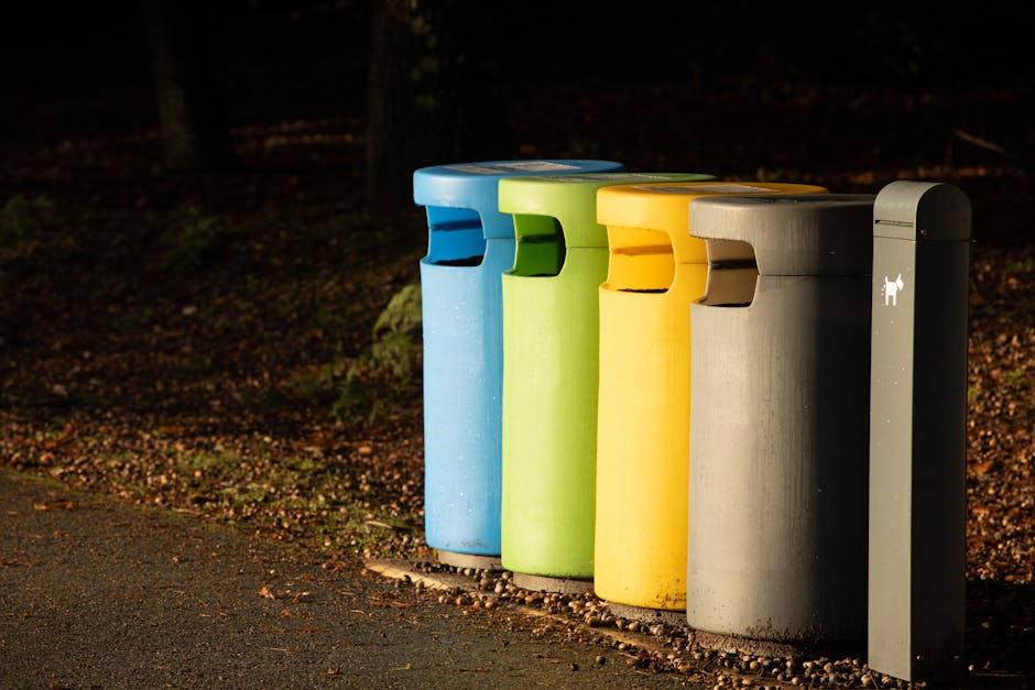 A row of four cylindrical, brightly colored waste bins positioned along a paved pathway, with the first bin in blue, the second in green, the third in yellow, and the fourth in gray. Each bin features a rectangular opening on the front for waste disposal, with a textured surface showing slight weathering. They are set on a small gravel border, adjacent to a natural, leaf-covered ground area with faintly visible trees and bushes in the background, suggesting an outdoor environment such as a park or residential street. To the right of the bins stands a slim, dark gray, upright waste disposal unit with a small icon of a dog, indicating it is for pet waste. The scene is lit with natural lighting, creating subtle shadows, and highlights the practical aspect of independent waste collection options, aligning with rubbish removal services in the local Dulwich area.