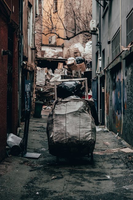 A narrow urban alleyway scene shows a large, grey, weathered refuse sack placed on a trolley or handcart, positioned in the foreground. The sack appears full and slightly crumpled, with visible creases and a rough, fabric-like texture. In the background, there is an overflowing pile of cardboard boxes, paper bags, and other waste materials stacked against a backdrop of aged brick and concrete buildings, some of which display graffiti. The alley has a dark, damp surface with patches of dirt, debris, and small puddles, indicating recent rain or cleaning. Metal pipes, electrical conduits, and a utility box are affixed to the building walls on either side, contributing to the urban atmosphere. Bare tree branches are visible above, suggesting winter or early spring. The lighting is diffuse, consistent with overcast weather, emphasizing the gritty, utilitarian environment often associated with private waste disposal or rubbish removal services such as those provided by Waste Clearance Dulwich, supporting the context of independent collection and on-site clearance in a typical city setting.