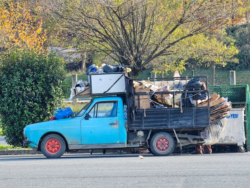 A large outdoor collection of mixed waste and rubbish is piled around a metal recycling bin on a paved street, situated near a parked silver car with a yellow license plate. The rubbish includes various cardboard boxes, plastic bags, paper, and packaging materials, some of which are torn or flattened. The recycling bin, marked for mixed paper and cardboard, is overflowing with discarded papers, boxes, and other waste materials spilling out. To the right, there are two black waste disposal bags and a bright red rubbish bin, all positioned on the curb, with additional cardboard and plastic waste scattered around the base. Behind the waste area, a low metal railing separates the refuse from the parking area, which also contains a tree with bare branches in the left foreground. The background features a commercial building with a storefront and a higher structure under construction or renovation, encased in blue scaffolding and mesh, exhibiting a typical urban streetscape. This scene illustrates a private waste collection scenario, potentially requiring independent rubbish removal services, such as those offered by Waste Clearance Dulwich, emphasizing the importance of proper waste disposal in a community setting.